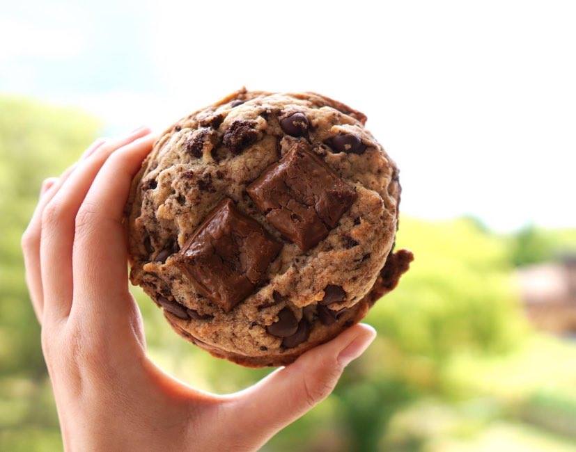 A hand holding up a chunky chocolate chip cookie with two Caramilk squares nestled ontop. Blurred, lush green background. 